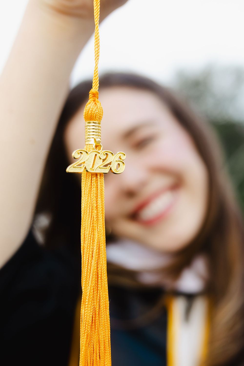 Smiling graduate holds up gold 2026 graduation tassel in focus against blurred background.