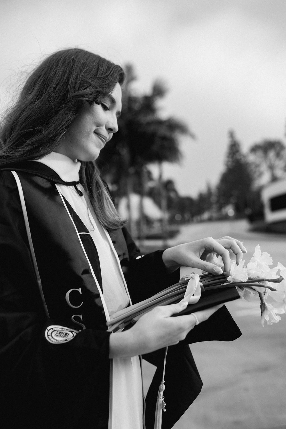 CSULB graduate in black gown holds diploma and white flower, smiling outdoors near palm trees.
