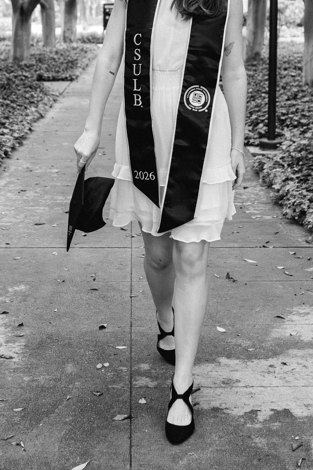 Black and white photo of a CSULB 2026 graduate in white dress holding cap, walking on campus sidewalk.