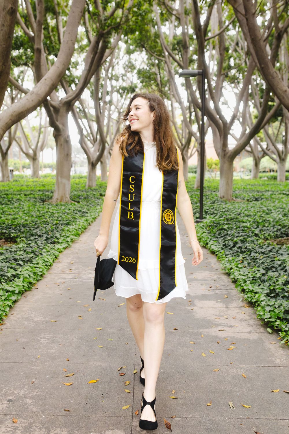 A smiling woman walks outdoors wearing a white dress and CSULB 2026 graduation stole, holding a cap.
