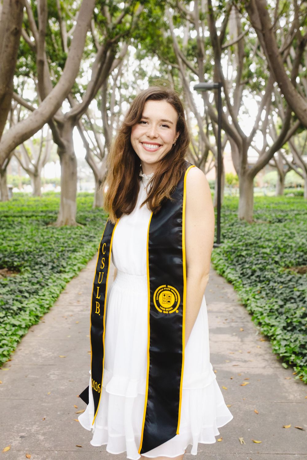 Smiling CSULB graduate wearing black and gold graduation stole over white dress, posing on tree-lined campus pathway.