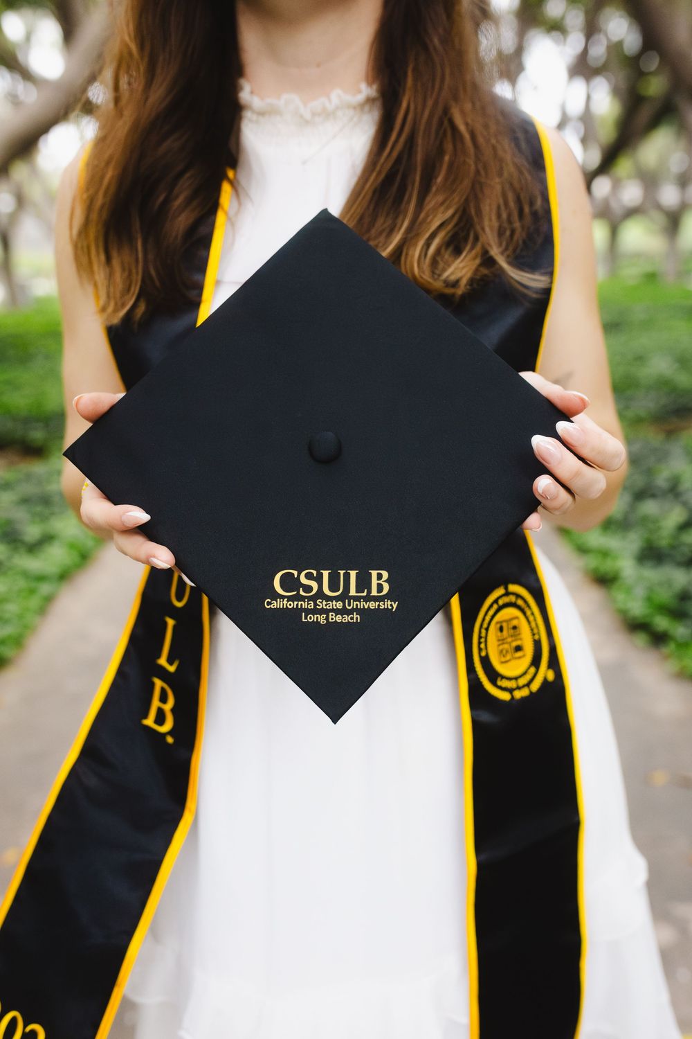 Graduate holding CSULB California State University Long Beach black mortarboard cap while wearing graduation stole.