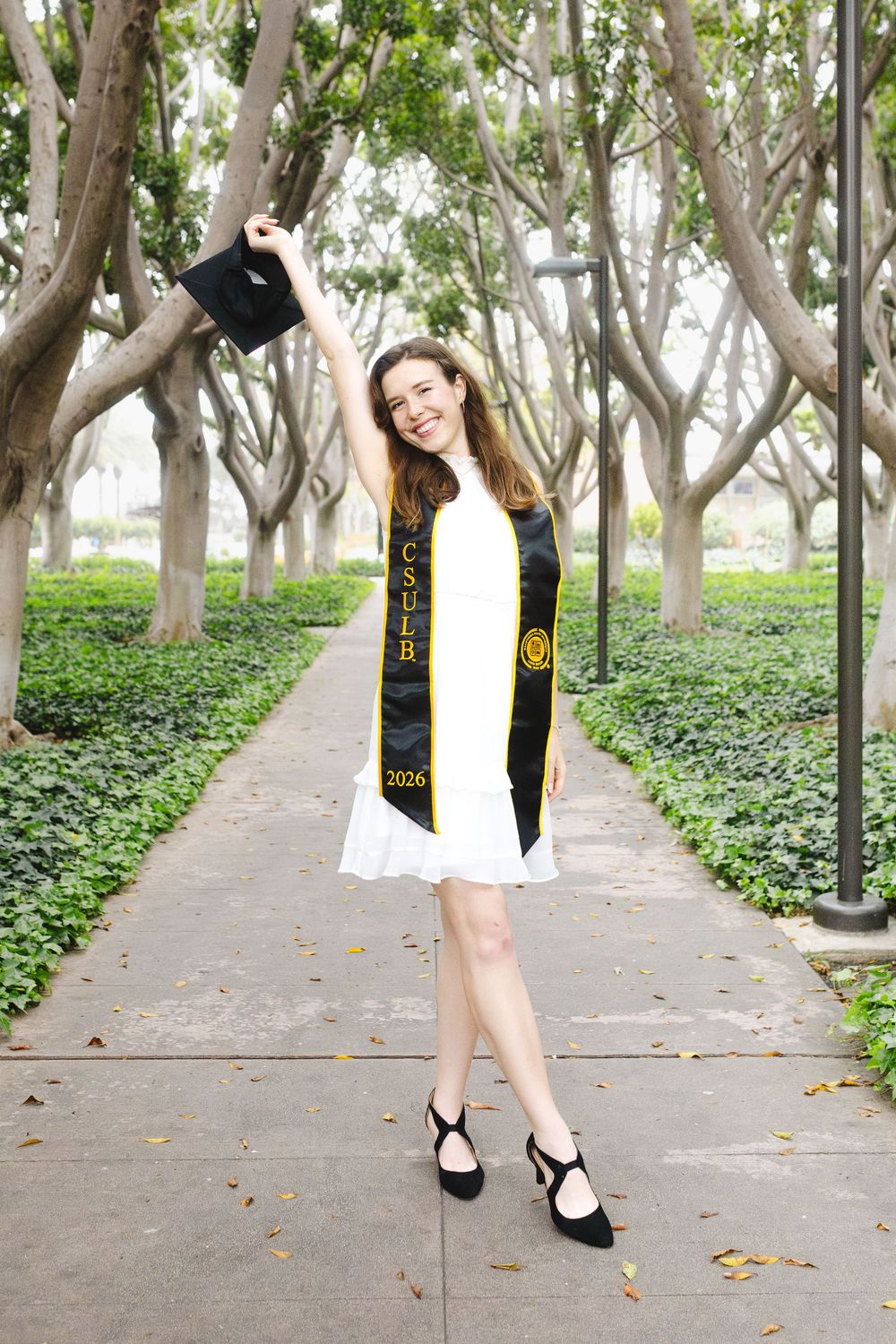 Smiling female CSULB 2026 graduate in white dress holding cap aloft on tree-lined campus walkway.