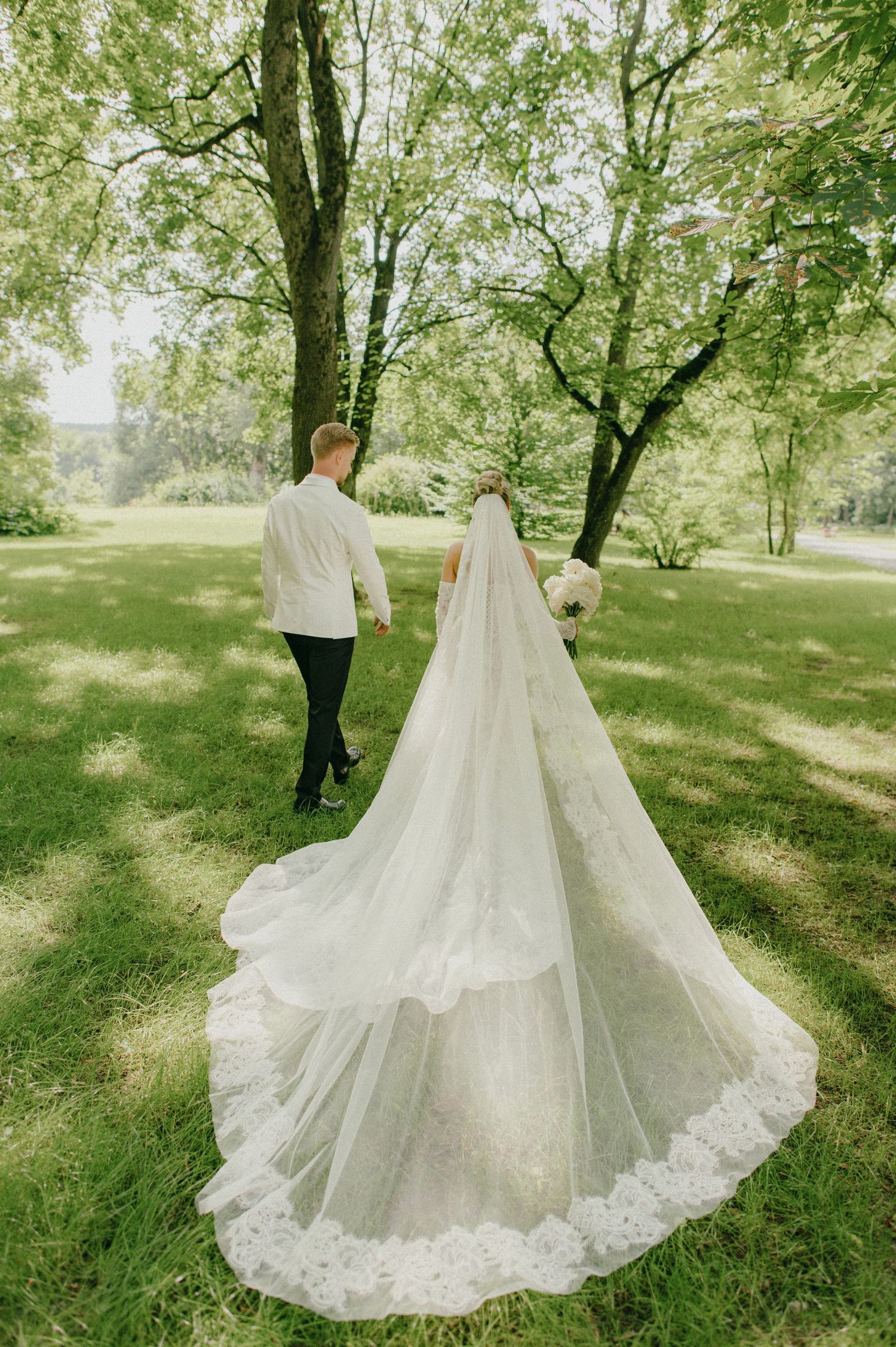Bride and groom walk hand-in-hand through lush green park, bride wearing long lace veil trailing on grass.