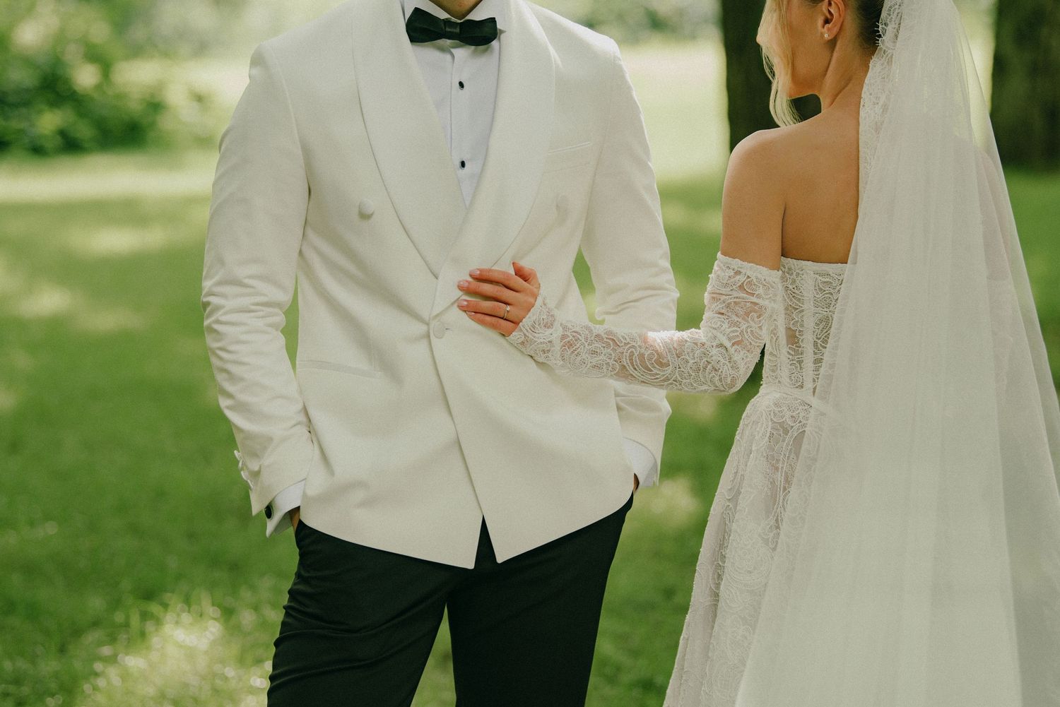 Bride and groom walking hand in hand outdoors, groom in white tuxedo jacket and bride in lace off-shoulder gown.