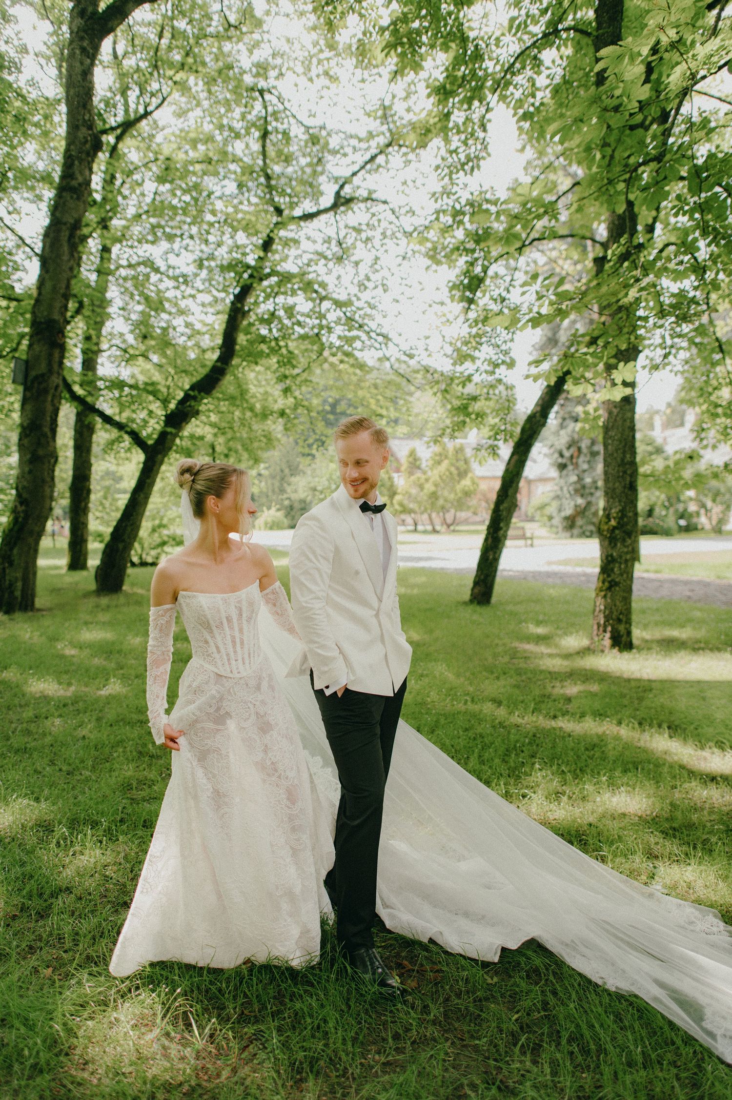 Bride in lace gown and groom in white tuxedo walk hand-in-hand through a lush green park.