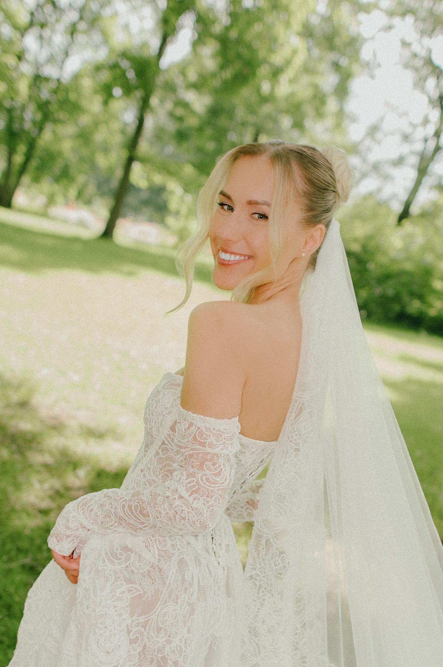 Smiling blonde bride in lace off-shoulder wedding dress with veil posing in a sunny green park.
