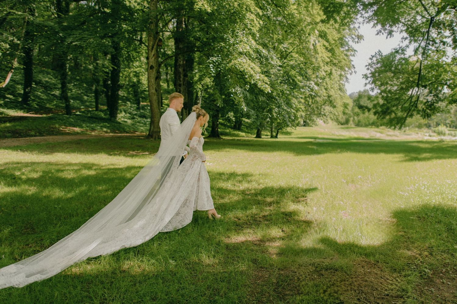 Wedding couple embrace in sunlit green meadow, bride in white gown with flowing cathedral veil.