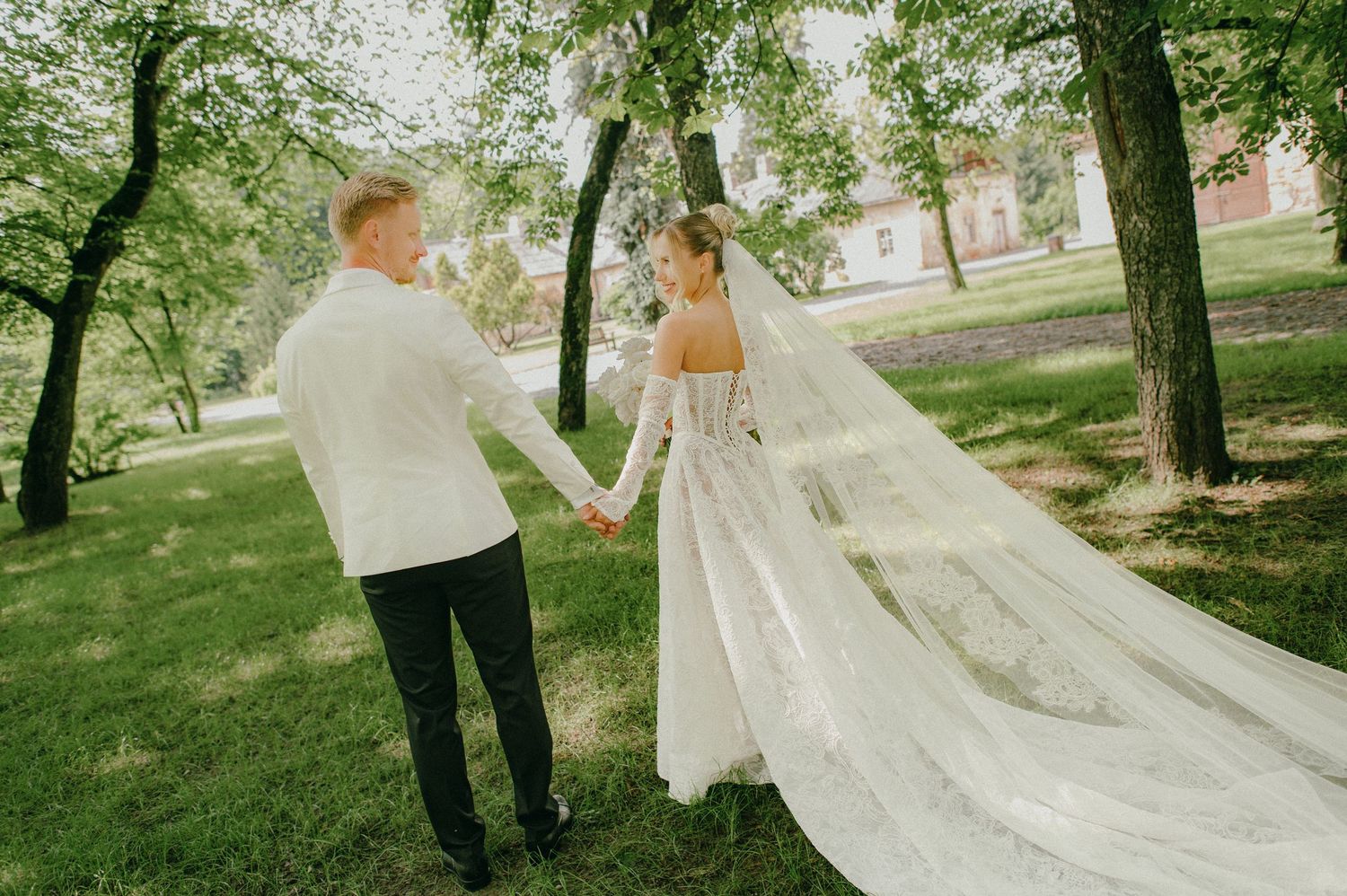 Bride and groom holding hands in a lush green park, bride wearing elegant white gown with long veil.
