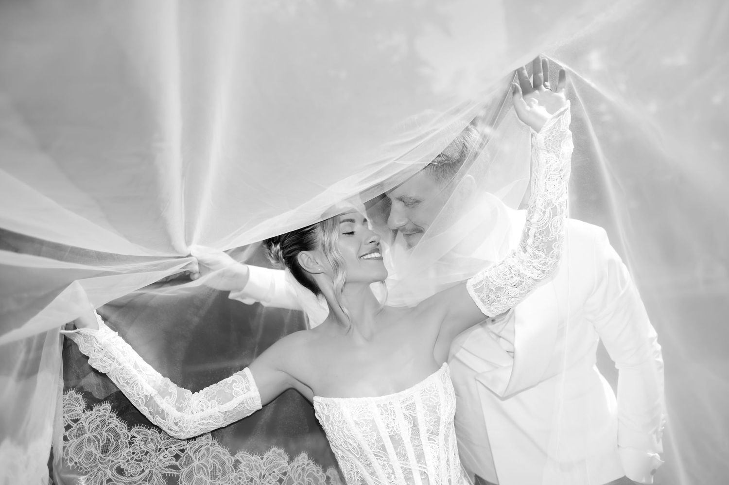 Bride in lace wedding dress smiling under flowing veil in elegant black and white wedding photo.