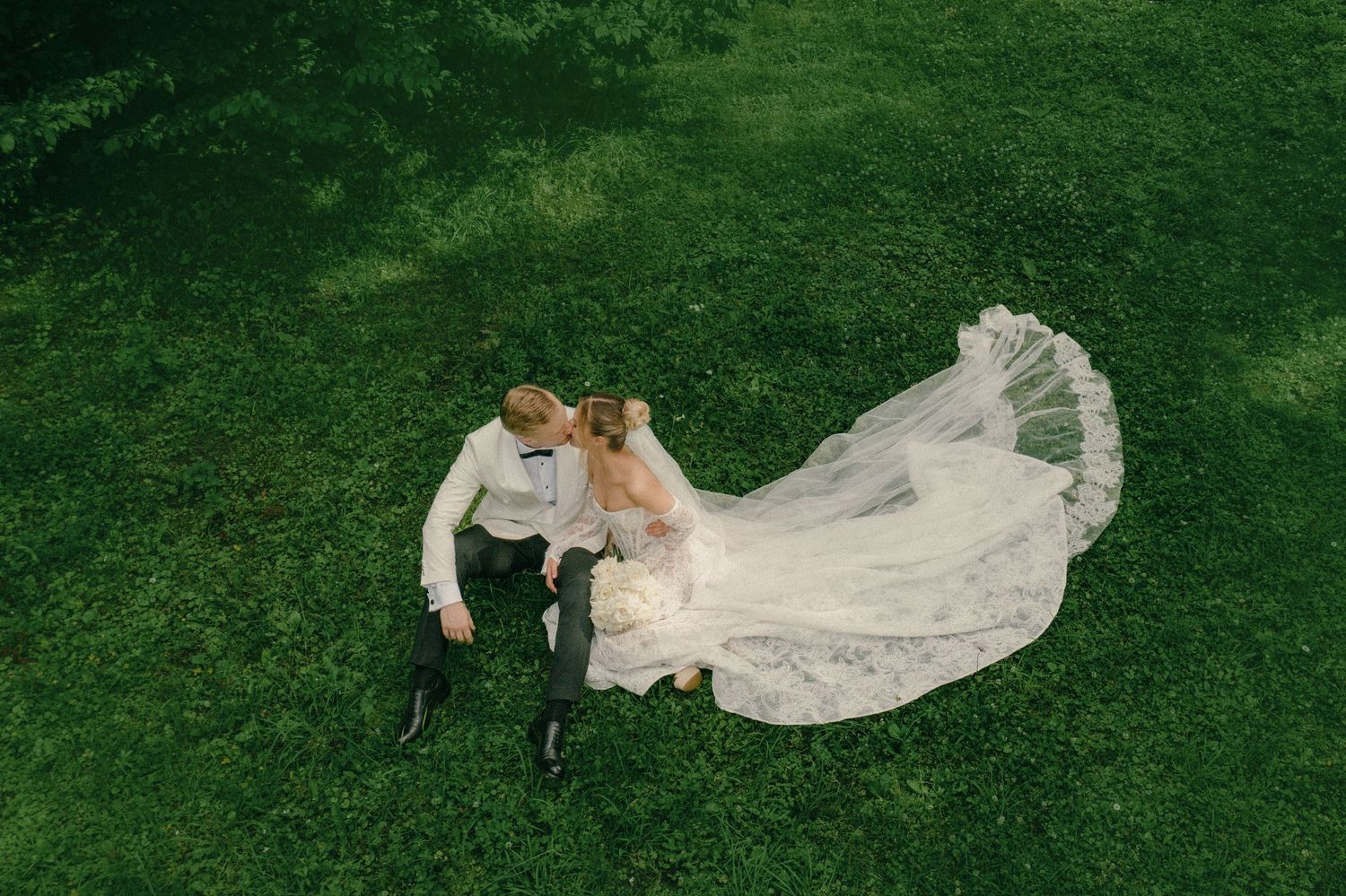 Aerial view of newlyweds sitting on green grass, bride in lace gown with flowing veil, groom in white shirt.