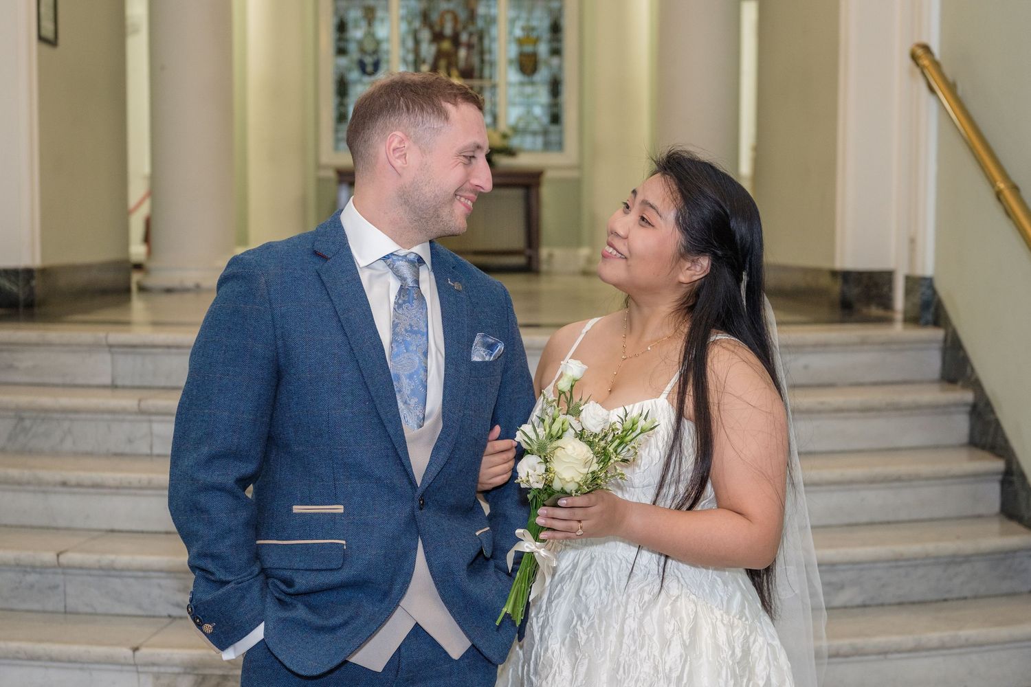 A newlywed couple in a church, groom in blue suit, bride in white gown holding bouquet, smiling at each other.