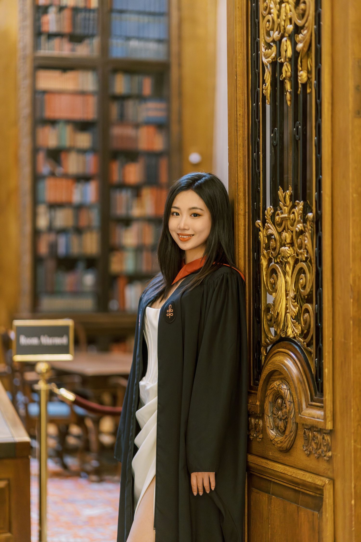 Young woman in Harvard graduation robes poses by ornate wooden door in a grand library with bookshelves.