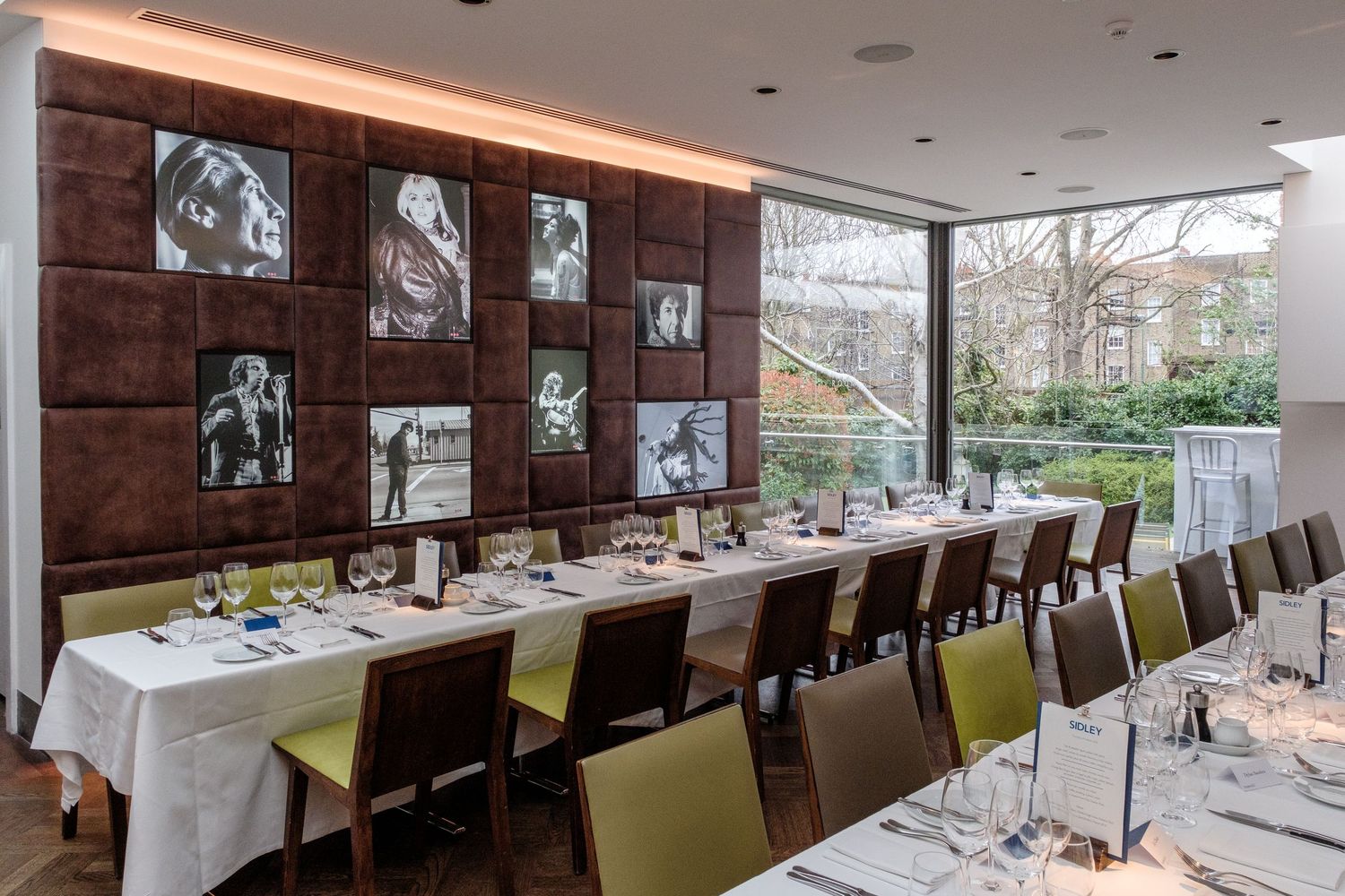Elegant private dining room with long white-clothed table, green chairs, black-and-white wall art, and floor-to-ceiling windows.