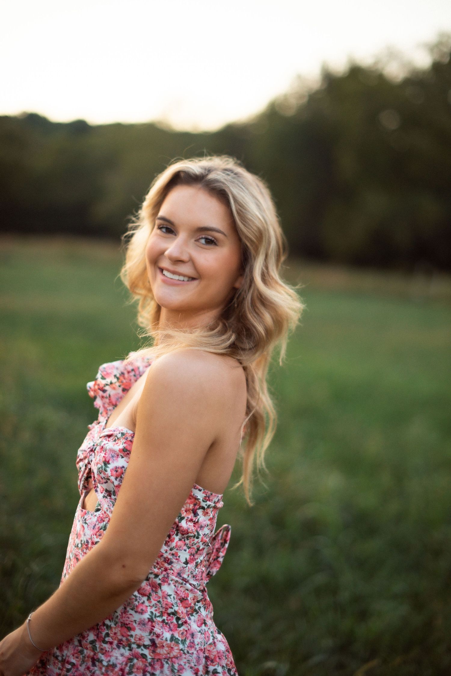 Young woman with long wavy blonde hair wearing a green dress and gold necklace smiling outdoors near a building.
