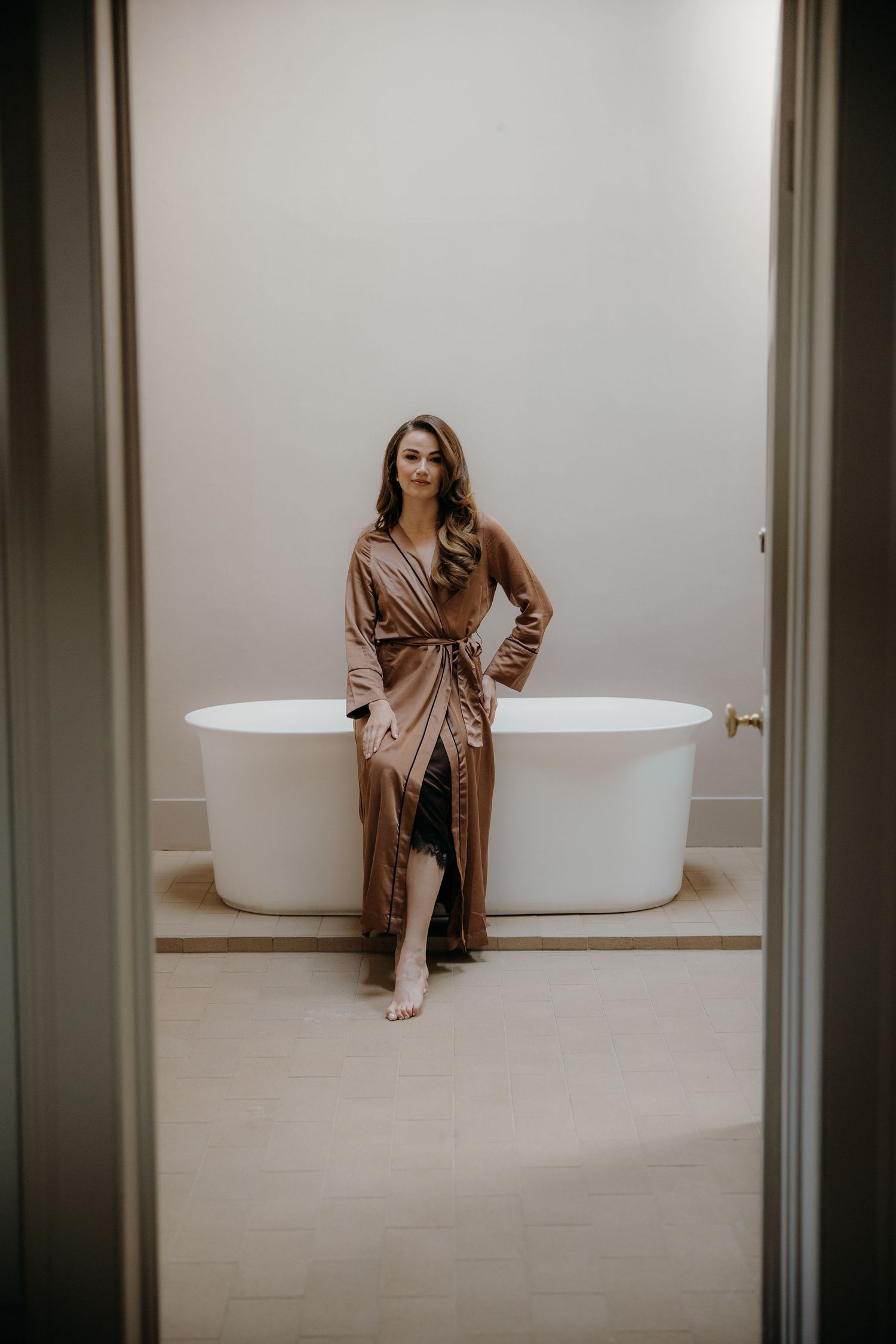 Woman in elegant bronze satin robe leaning against freestanding white bathtub in luxury bathroom.