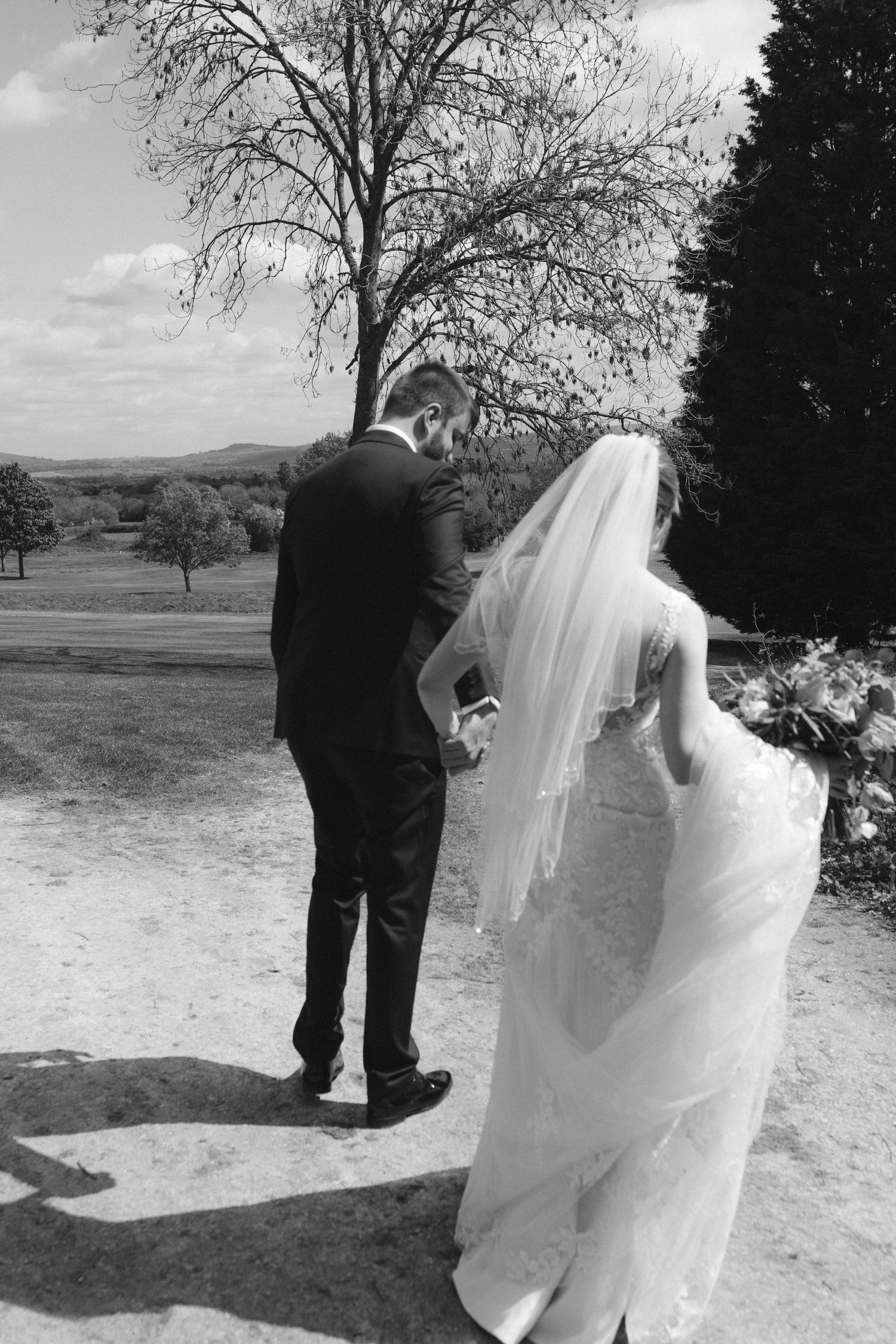 Black and white photo of bride and groom holding hands, walking through scenic outdoor wedding venue with trees.