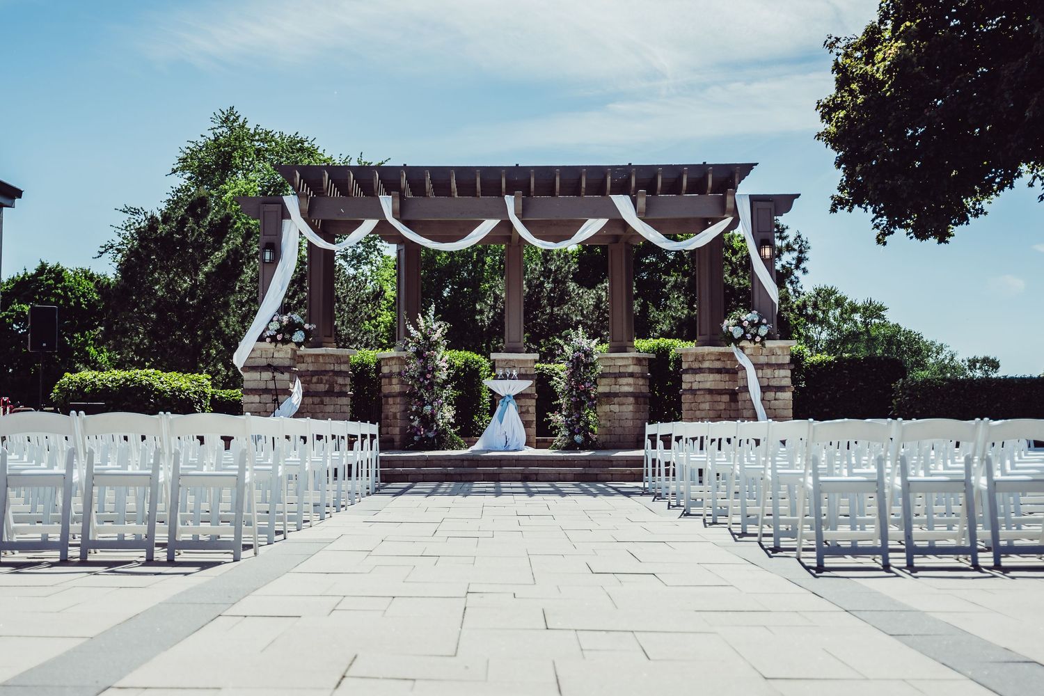 Elegant outdoor wedding ceremony setup with white chairs, floral arch, and stone pavilion under a bright blue sky.