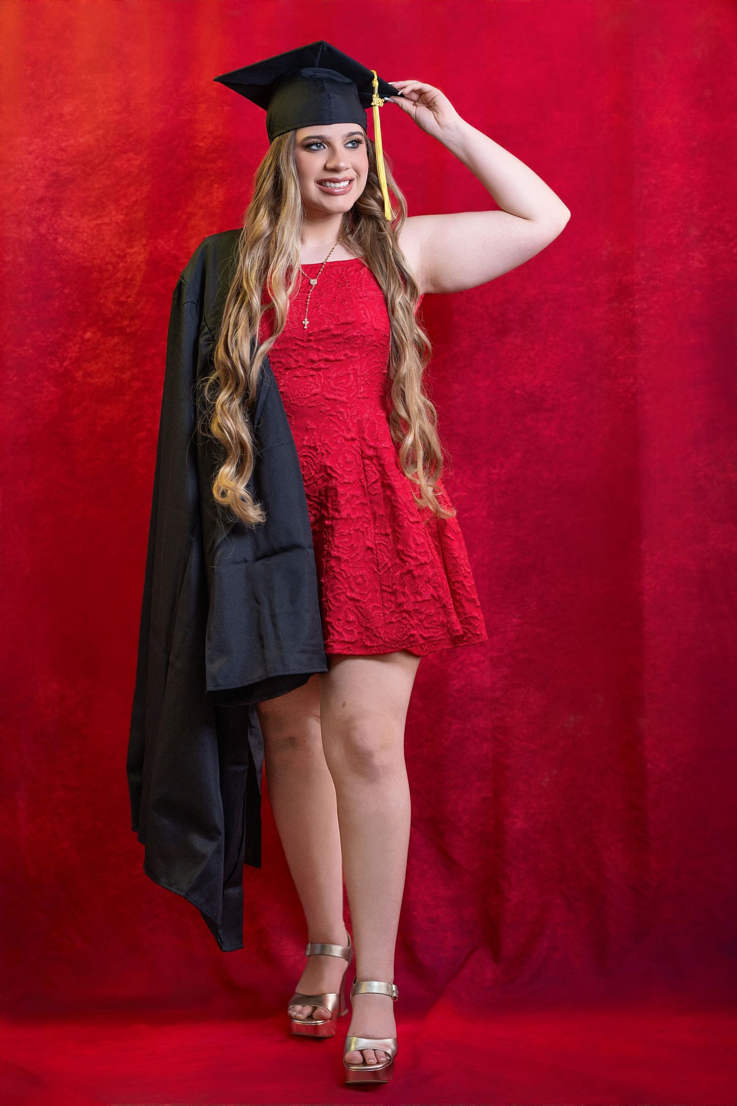 Young woman in red lace dress and graduation cap and gown posing against red background.