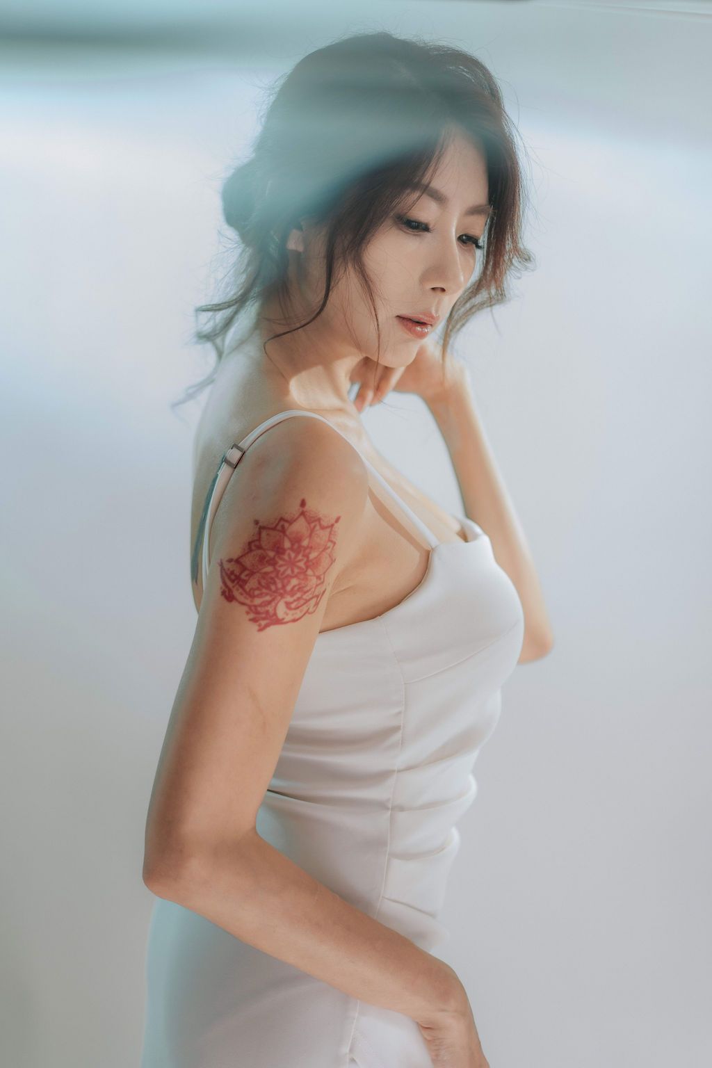 Young Asian woman in white dress with red floral tattoo on shoulder, posing elegantly against bright background.
