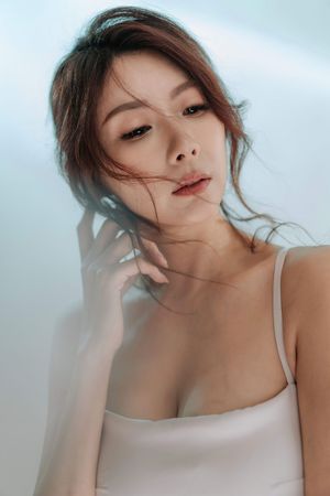 Young Asian woman in white tank top posing gracefully with hand near face against soft blue background.