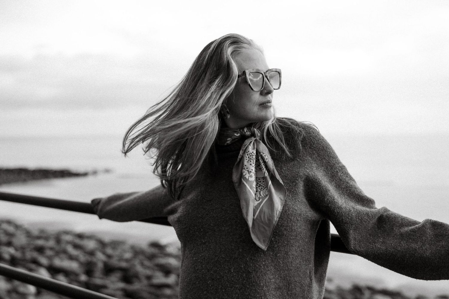 Black and white photo of a woman with windswept hair wearing glasses and a scarf by the seaside.