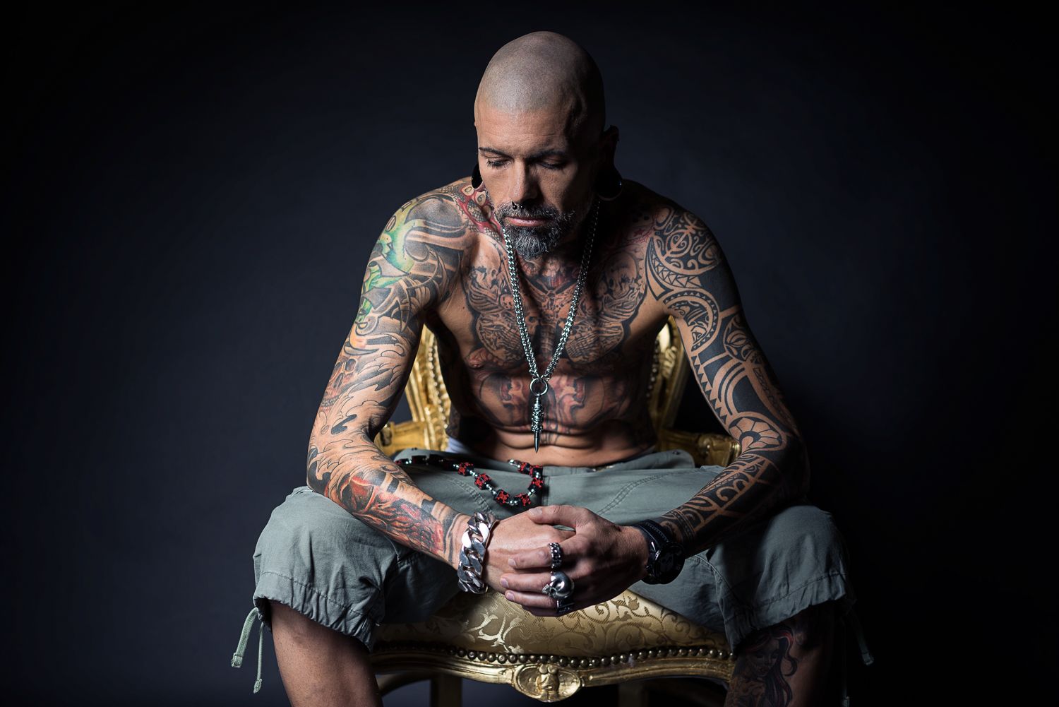 Heavily tattooed bald man sitting on ornate chair in dramatic studio portrait with dark background.