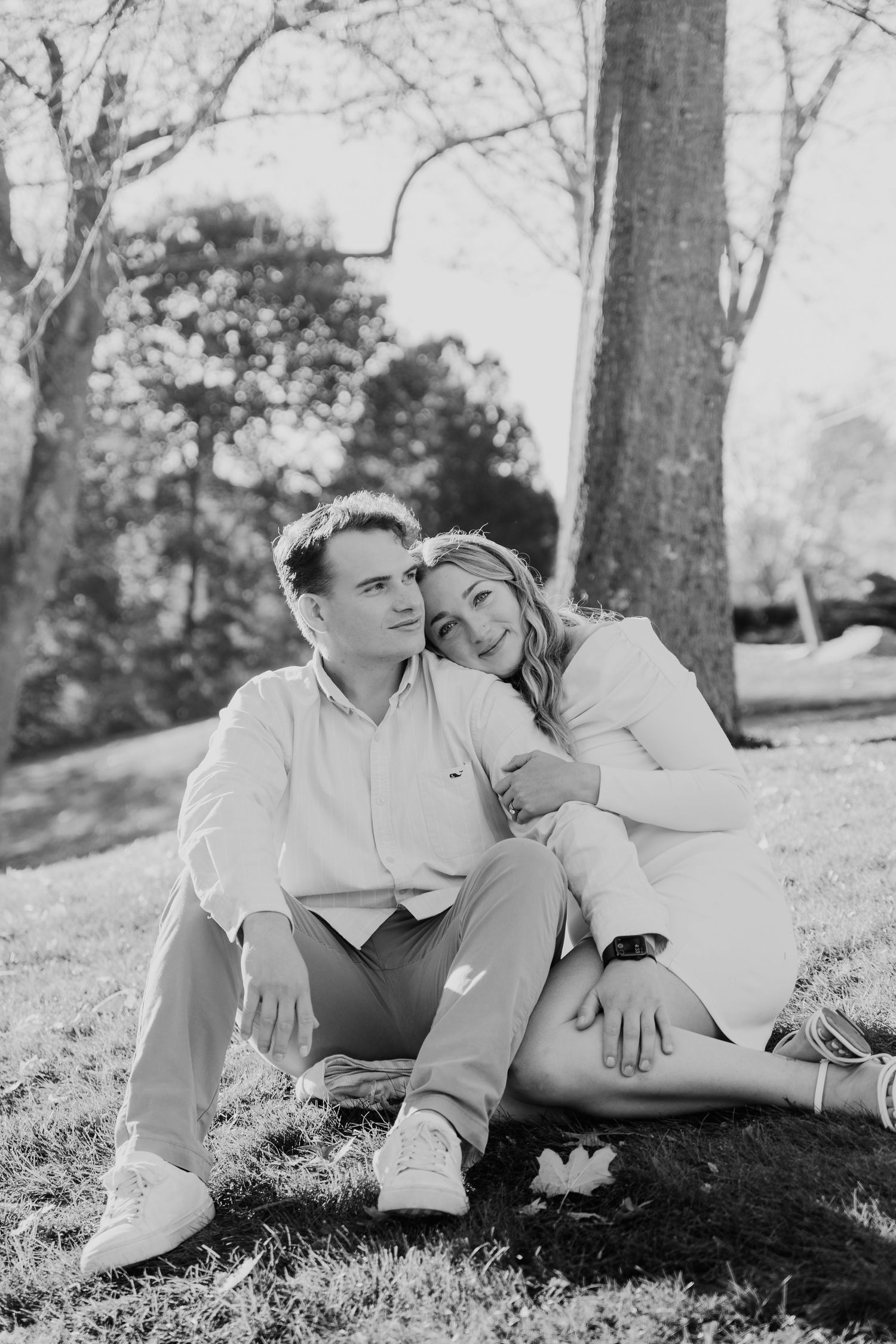 Black and white photo of a couple sitting on grass in a park, woman resting her head on mans shoulder.