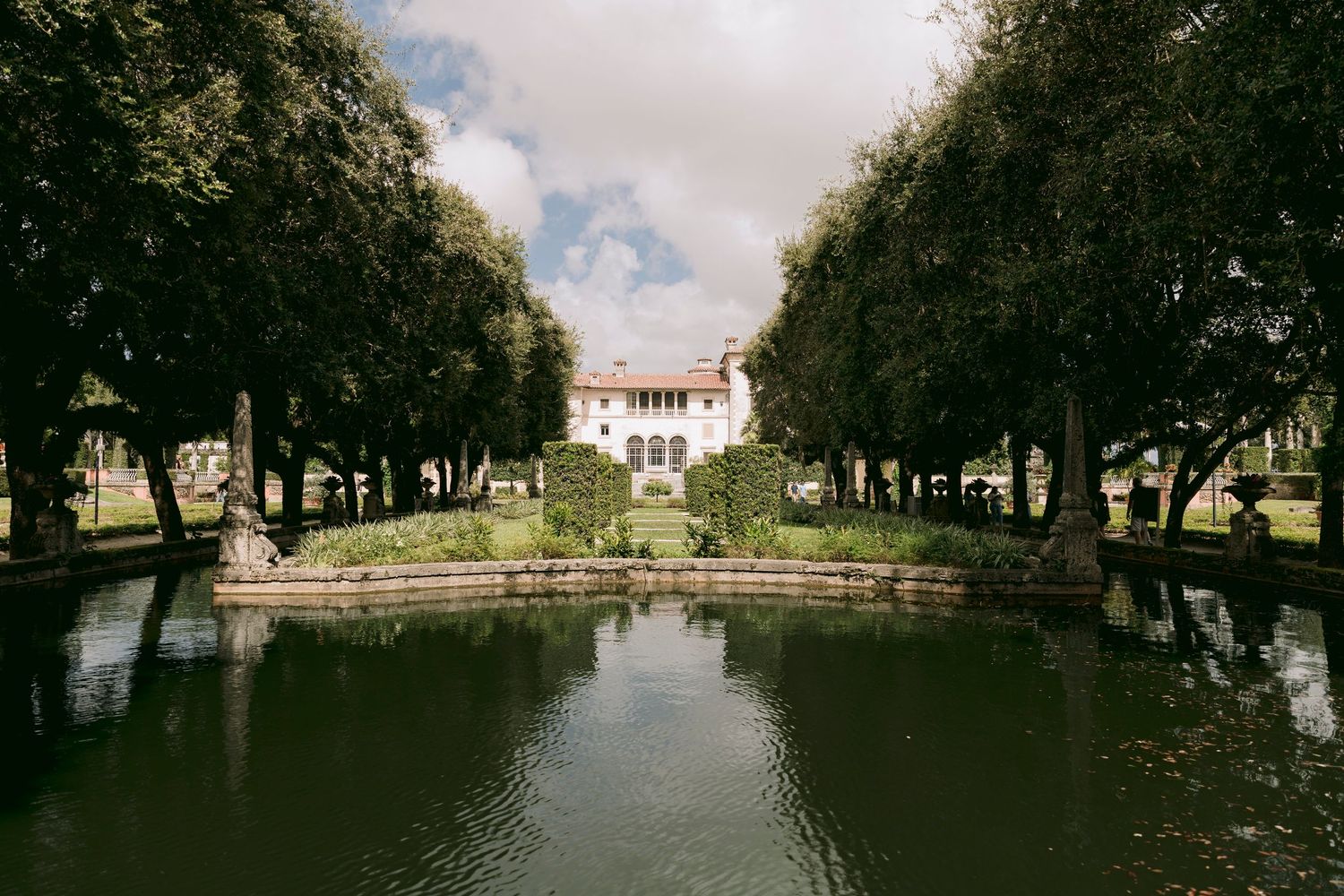 Grand white villa framed by tall trees reflects in a serene canal at Vizcaya Museum and Gardens, Miami.