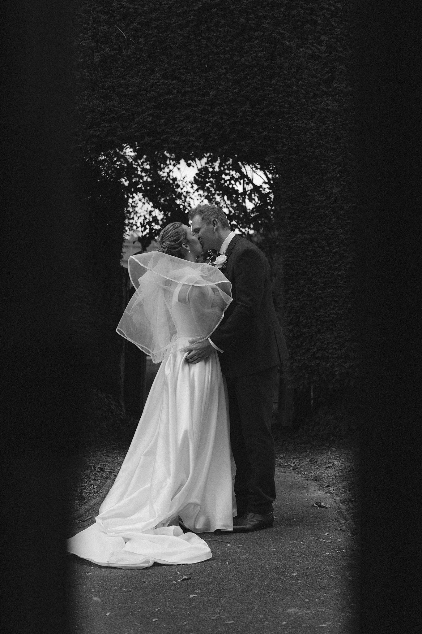Bride in white gown and veil walks with groom in black suit through a churchyard cemetery on their wedding day.