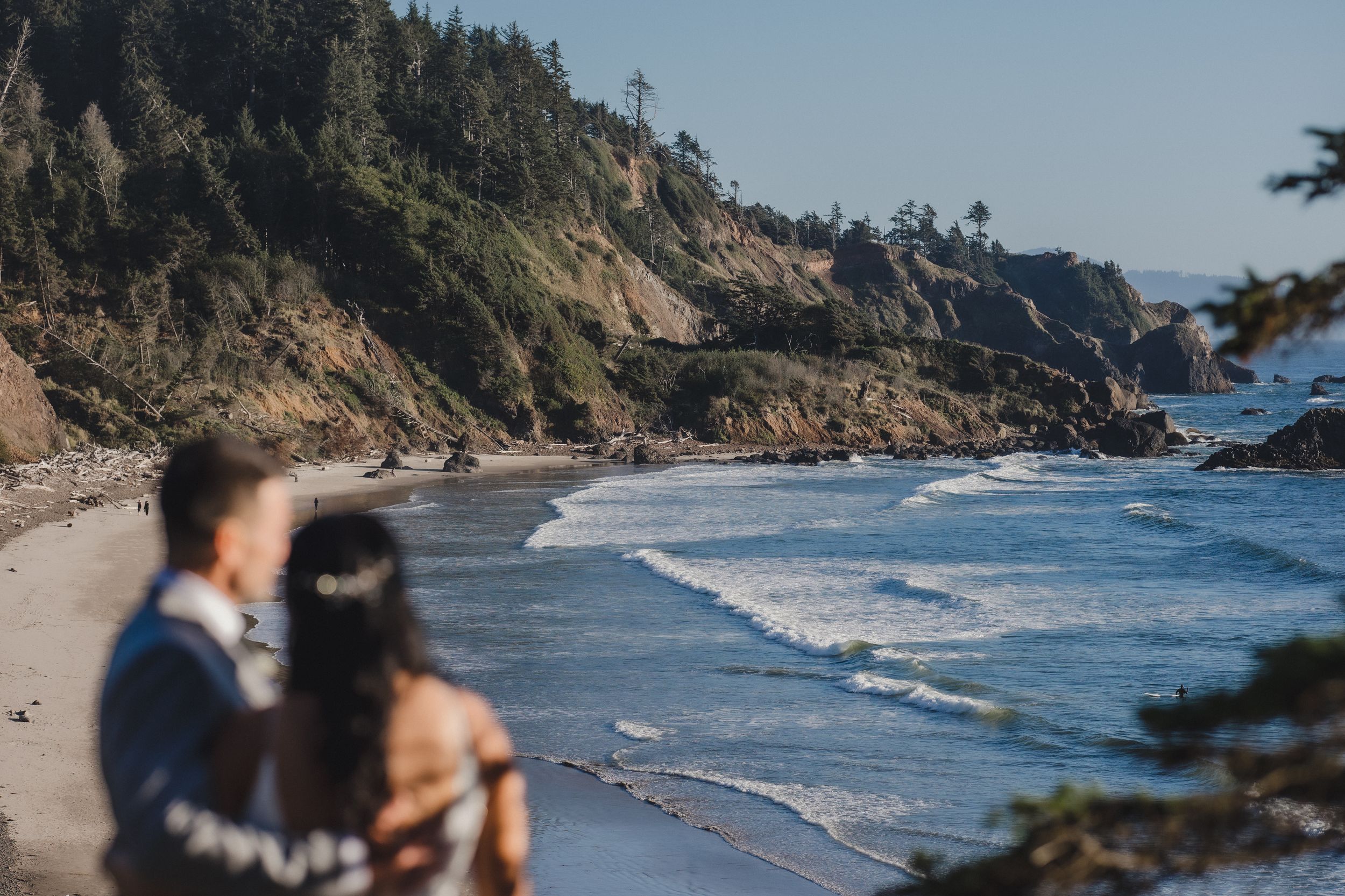 Couple stands on a scenic coastal beach with rocky cliffs, waves, and lush trees on a sunny day.