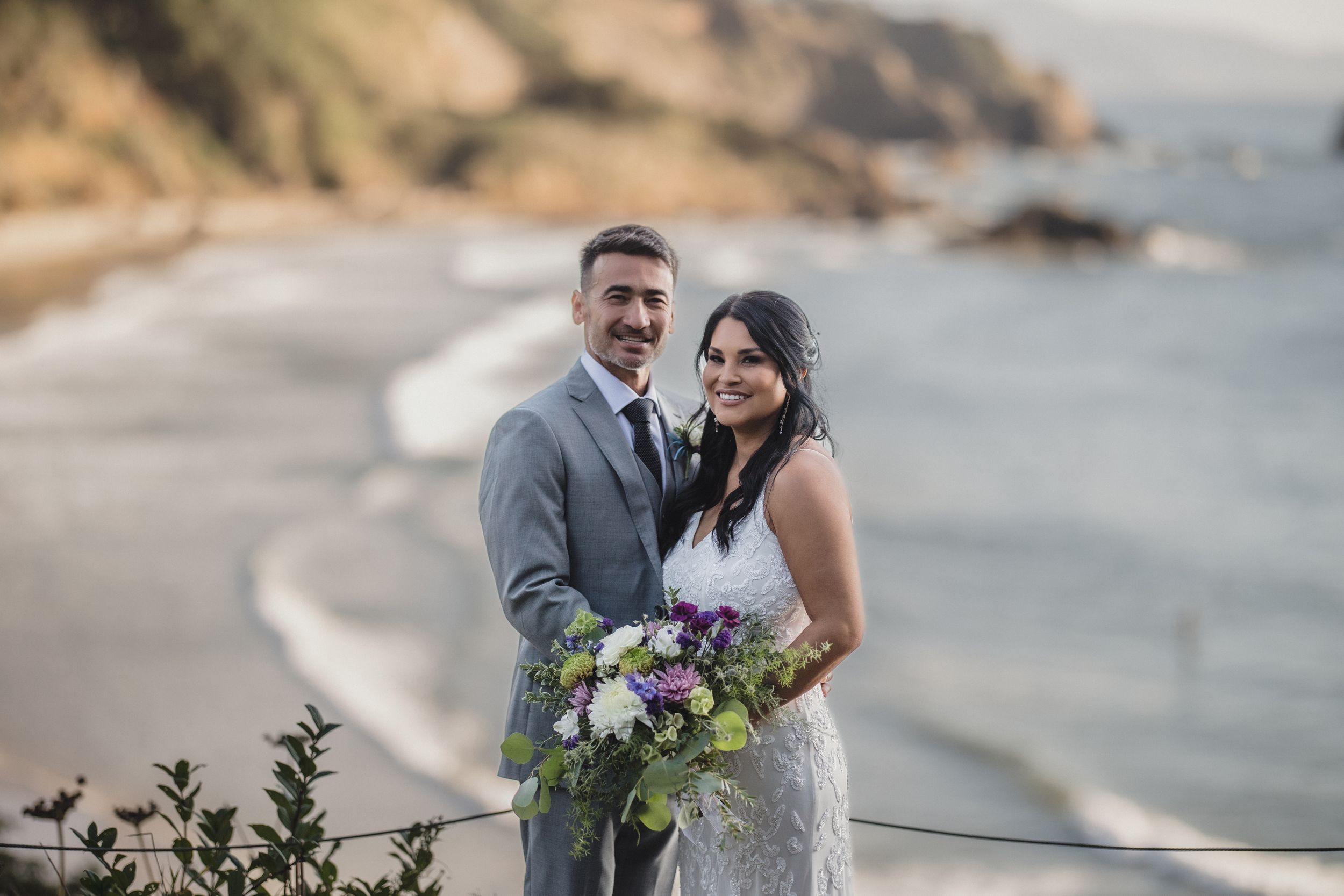 Couple in wedding attire smiling on coastal cliffside, bride holding purple bouquet with ocean backdrop.