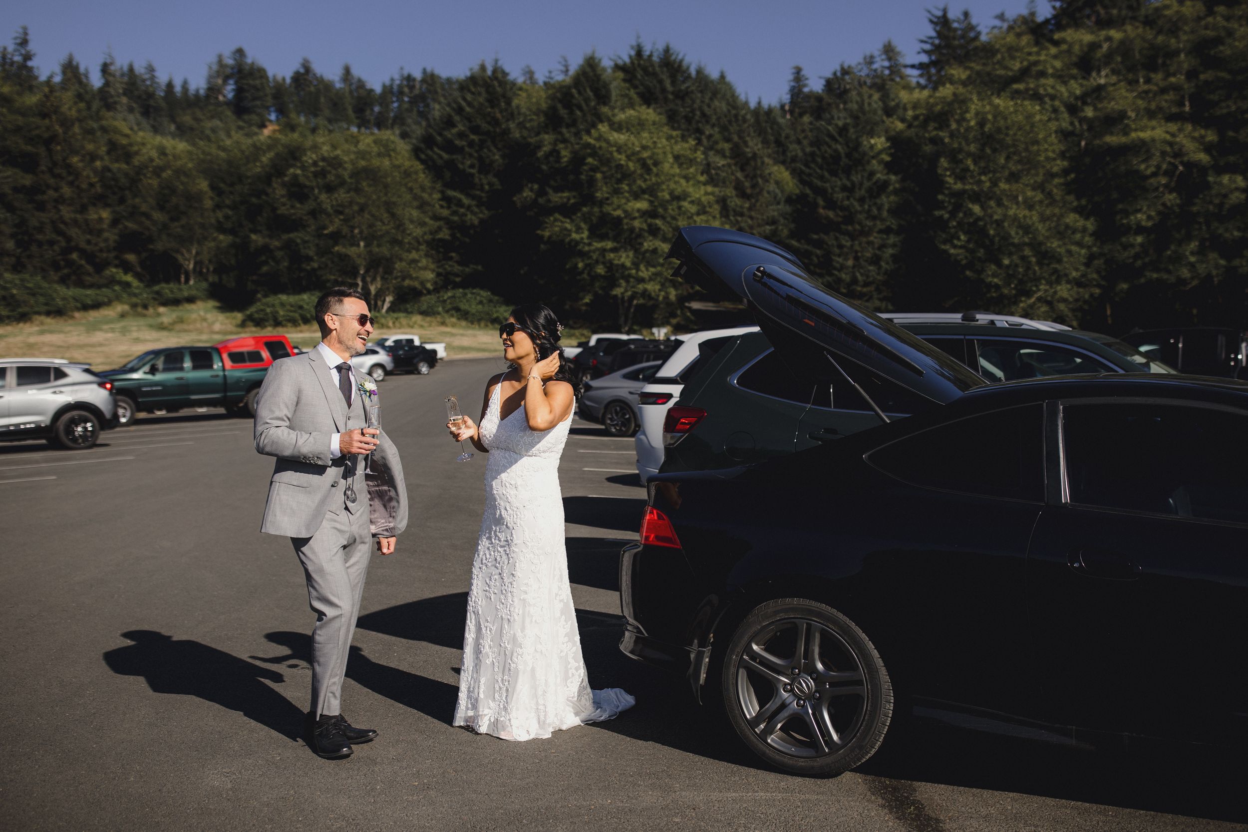 Bride and groom in wedding attire standing beside a black SUV in a parking lot surrounded by evergreen trees.