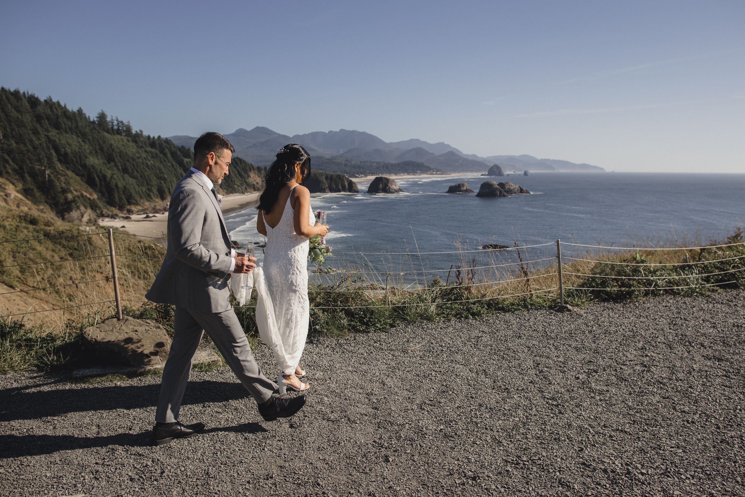A bride and groom walk hand in hand along a scenic coastal overlook with ocean views and rock formations in the background.