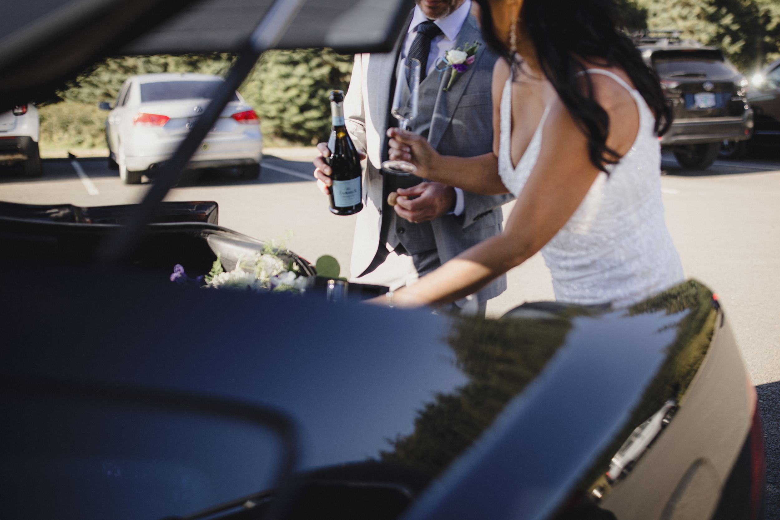 Bride and groom opening champagne at car trunk on wedding day.