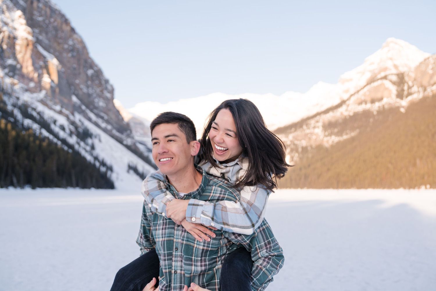 A happy couple enjoys a piggyback ride on a frozen Lake Louise, Banff with snow-capped mountains in the background.