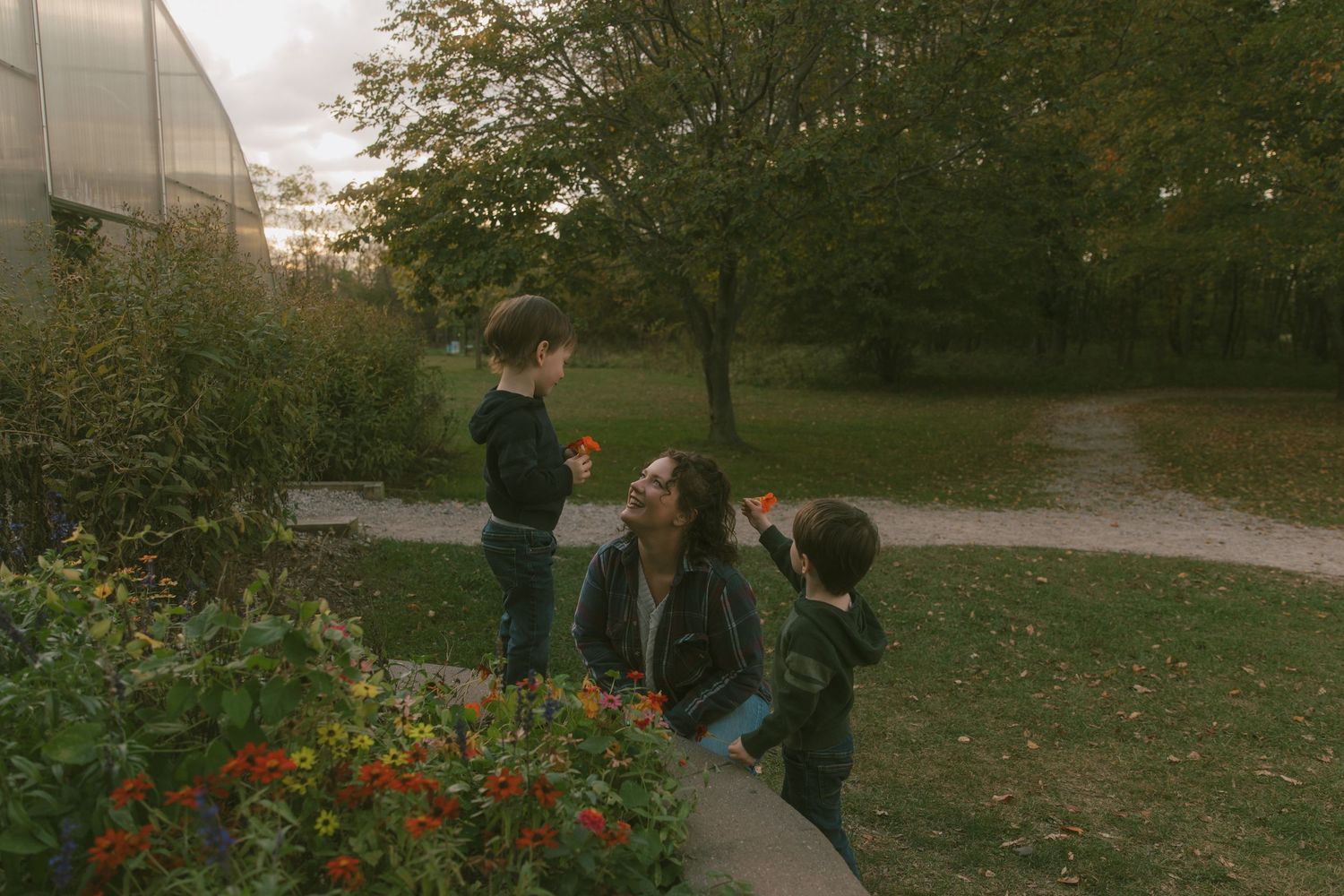 A mother crouches beside two children admiring colorful flowers in a lush garden near a greenhouse on a cloudy day.