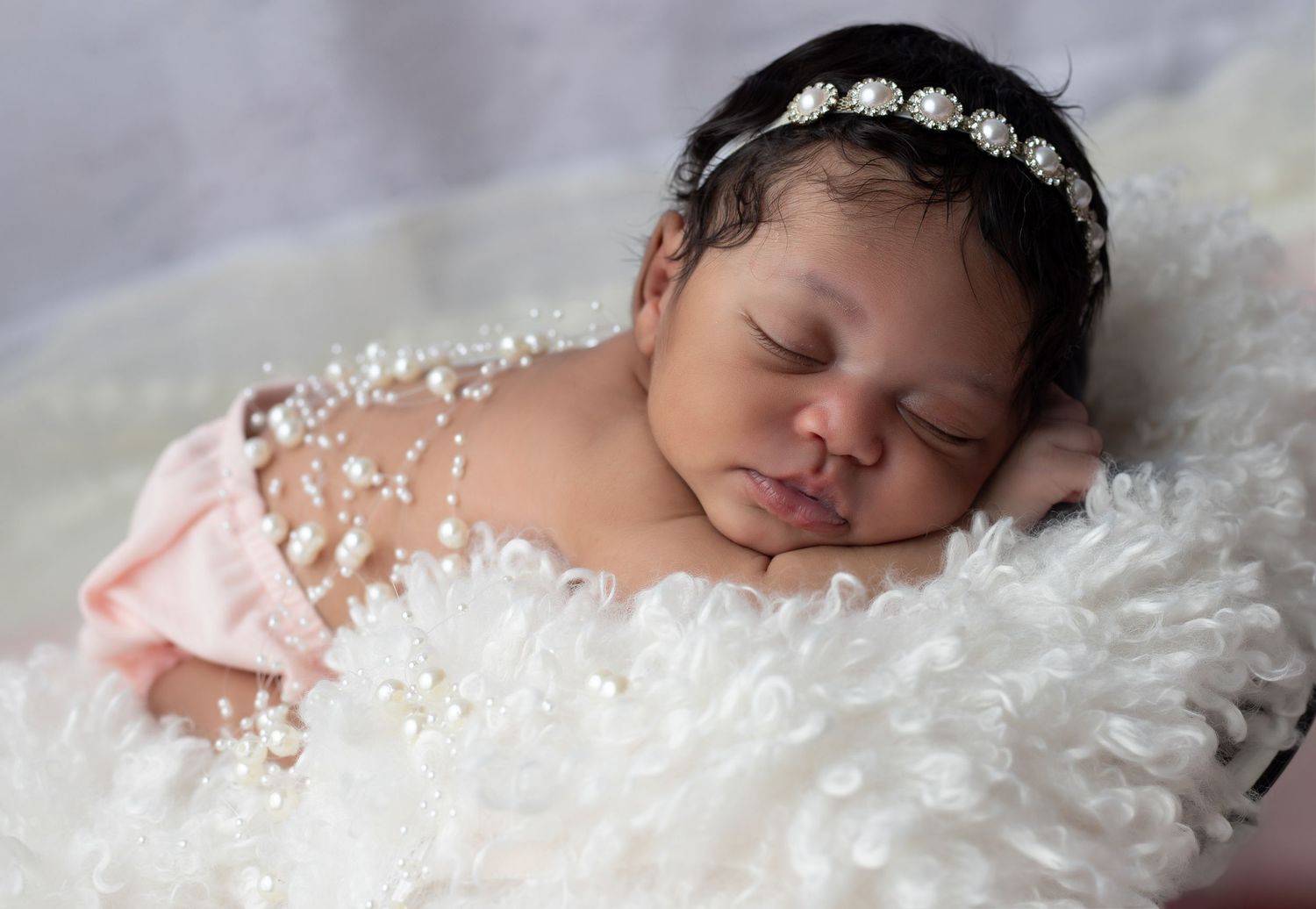 Sleeping newborn baby girl laying on white fluffy fabric and pearls on her back with little pink diaper cover.