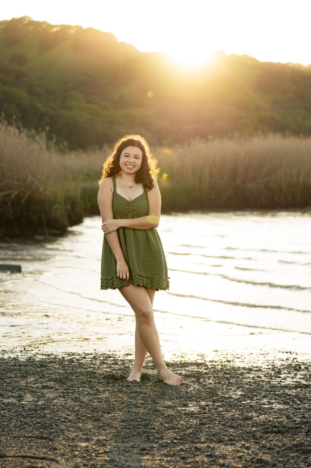 Person in olive dress poses playfully on beach during golden hour.