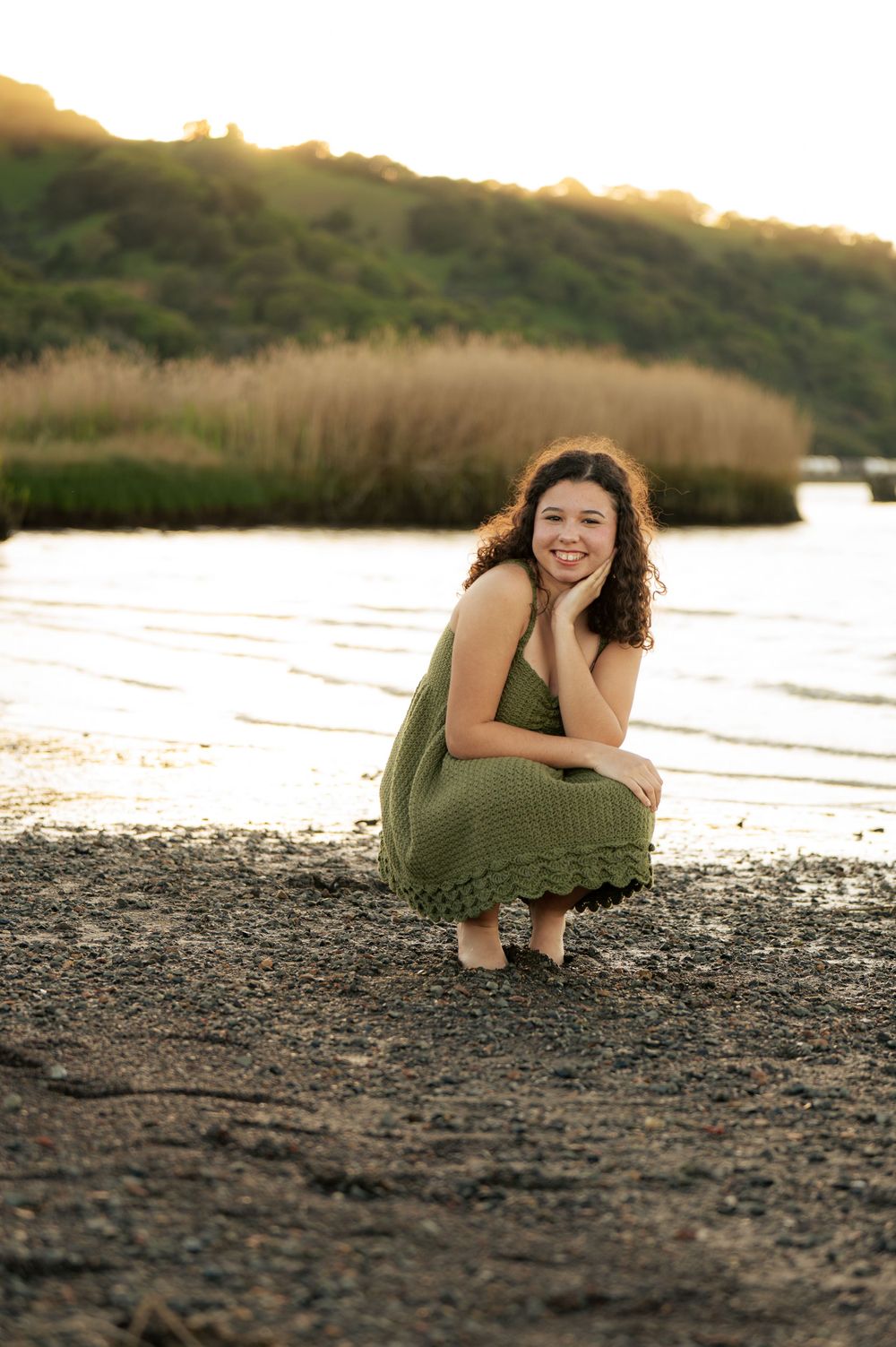 A person in a green dress poses playfully on a rocky beach at sunset.