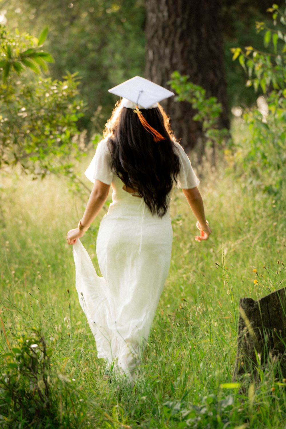 Graduate in flowing white dress twirling in sunlit garden setting.