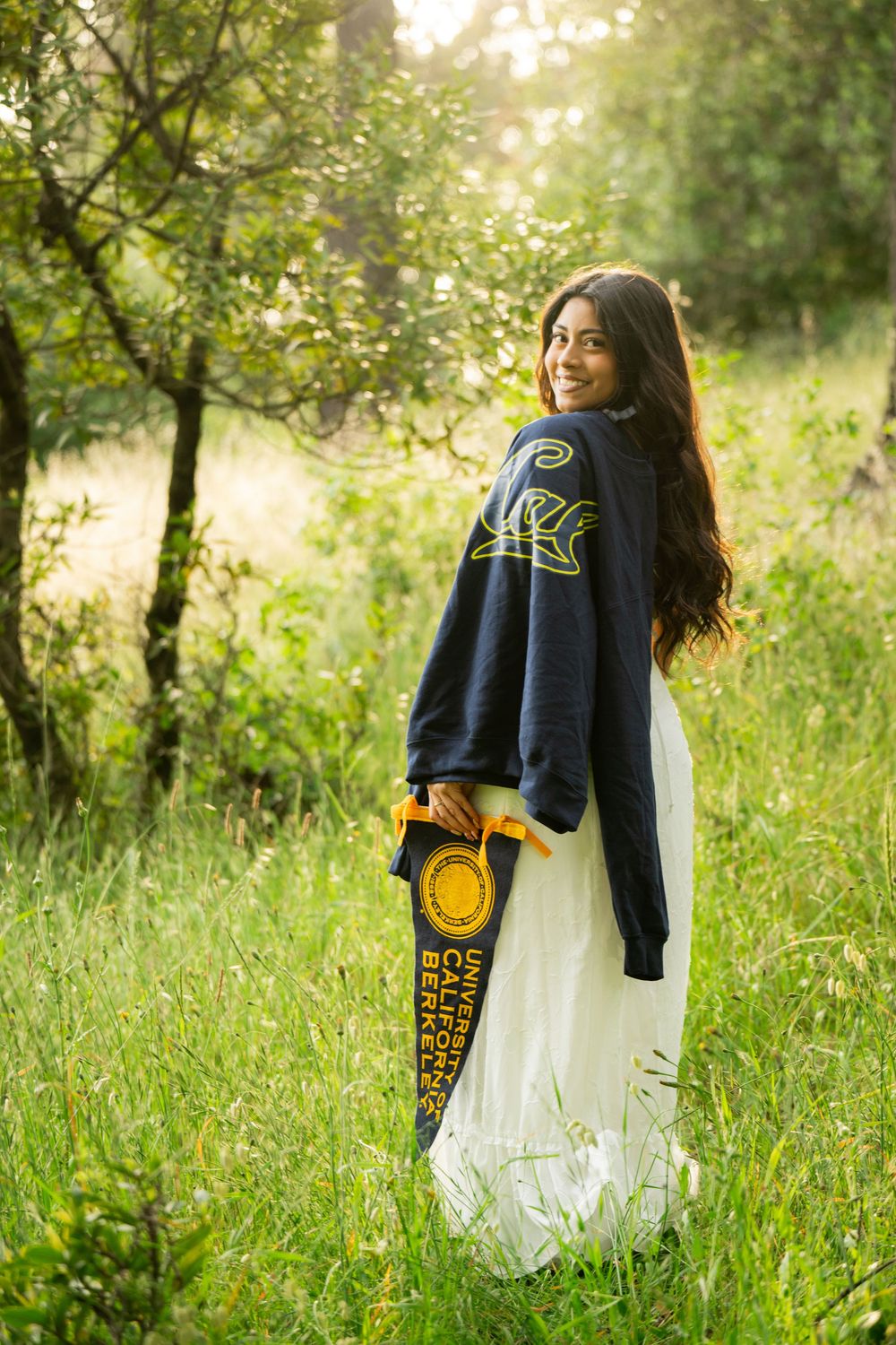 Graduate in white dress and navy varsity jacket poses in a sunlit grassy field.