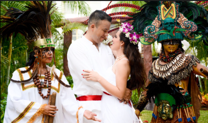 Couple shares romantic kiss at Mayan wedding ceremony with costumed performers in colorful traditional attire outdoors.
