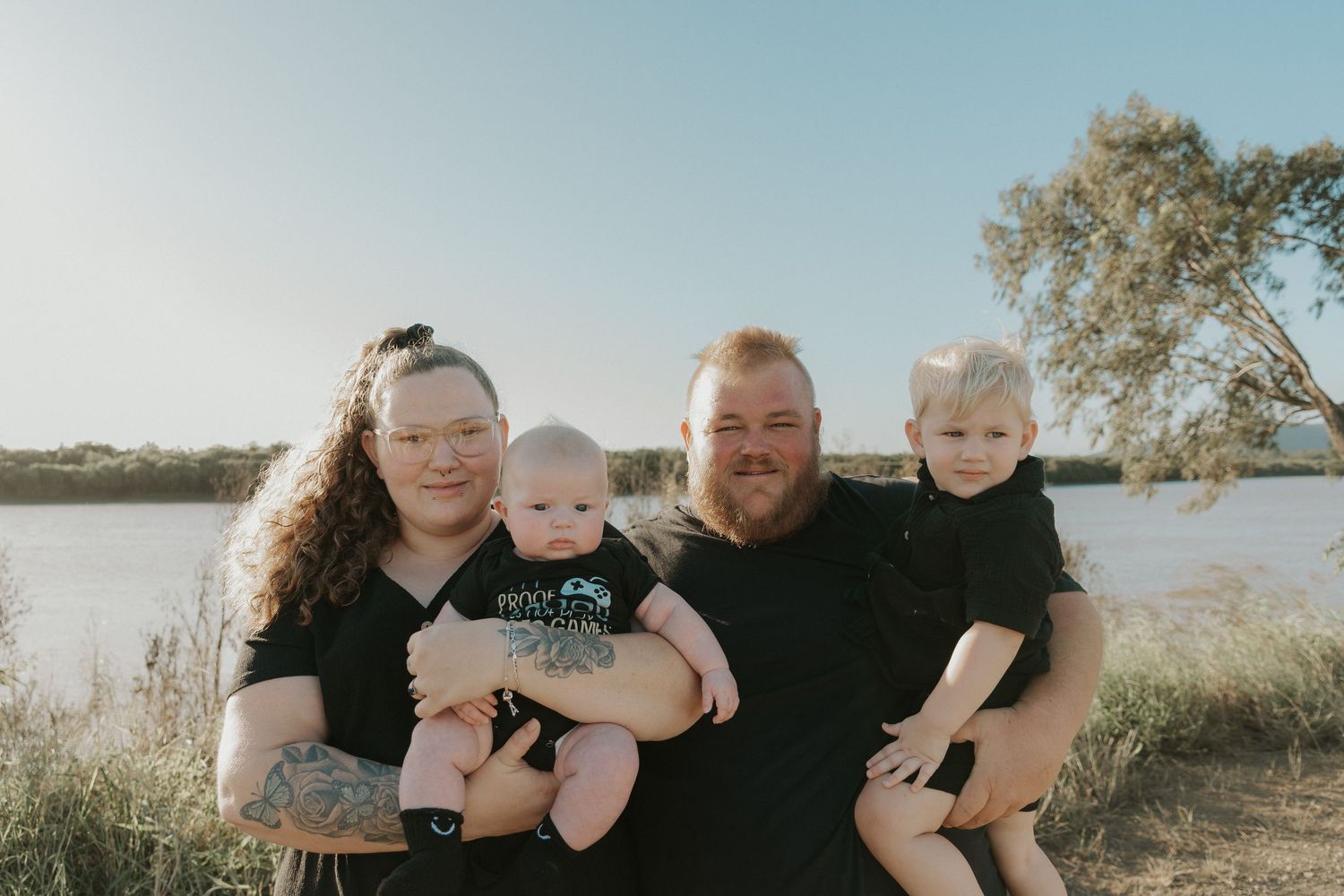 Happy family of four posing outdoors near a river, parents holding two young children on a sunny day.