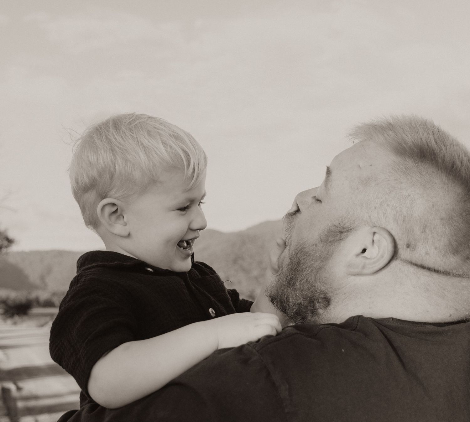 Bearded father sharing a playful nose-to-nose moment with his laughing young son in black and white.