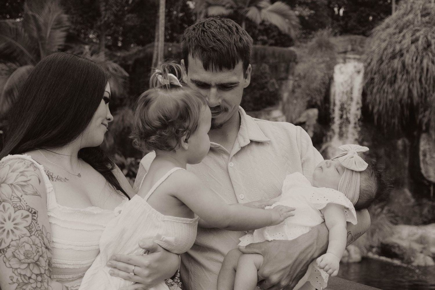 Black and white family portrait of parents with toddler and newborn near a garden waterfall.