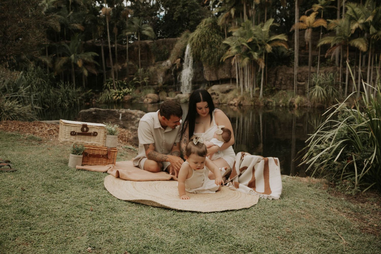 Young family enjoying a picnic on a blanket near a tropical waterfall with their two children.