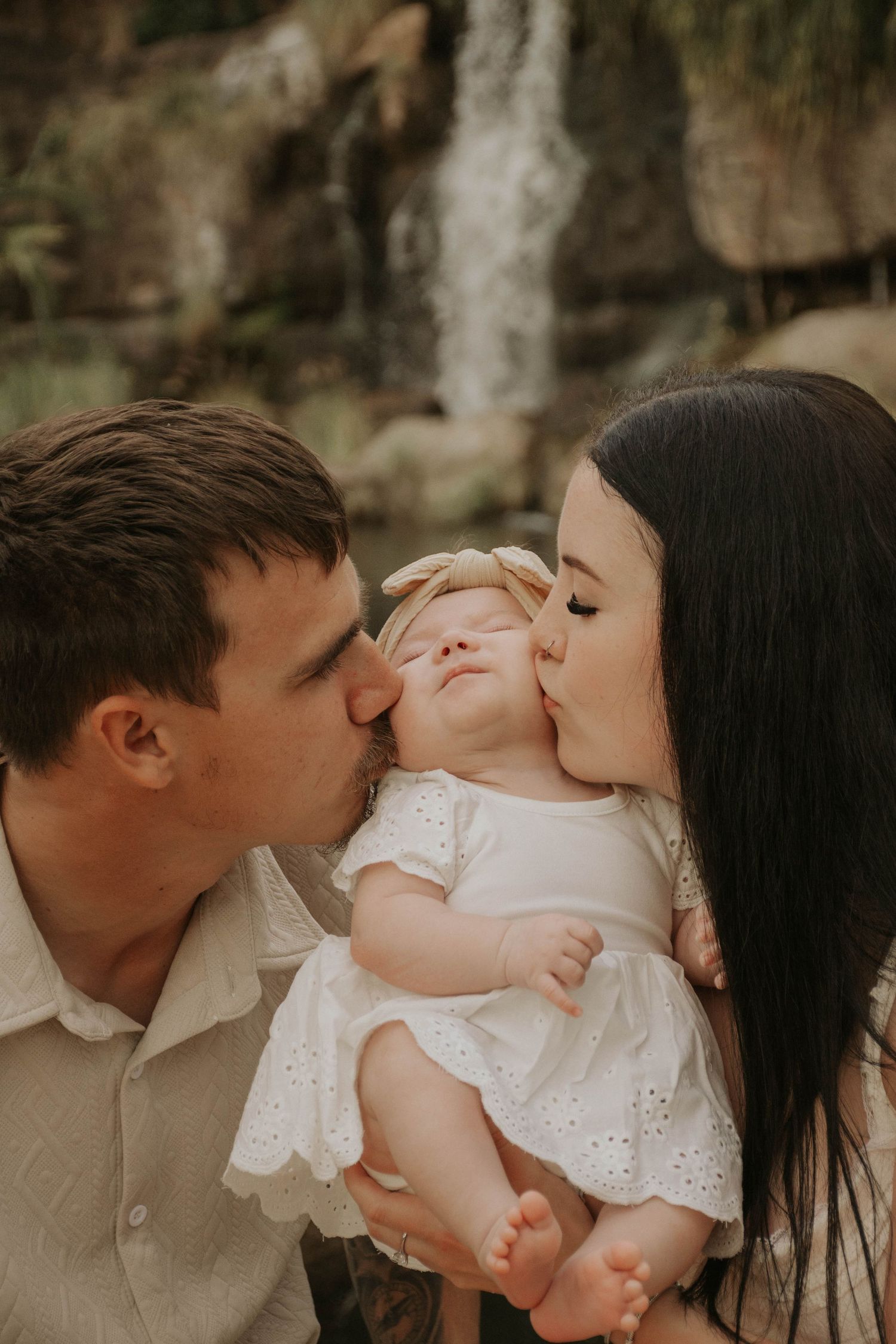 Loving parents simultaneously kissing their newborn baby girl on the cheeks near a waterfall.