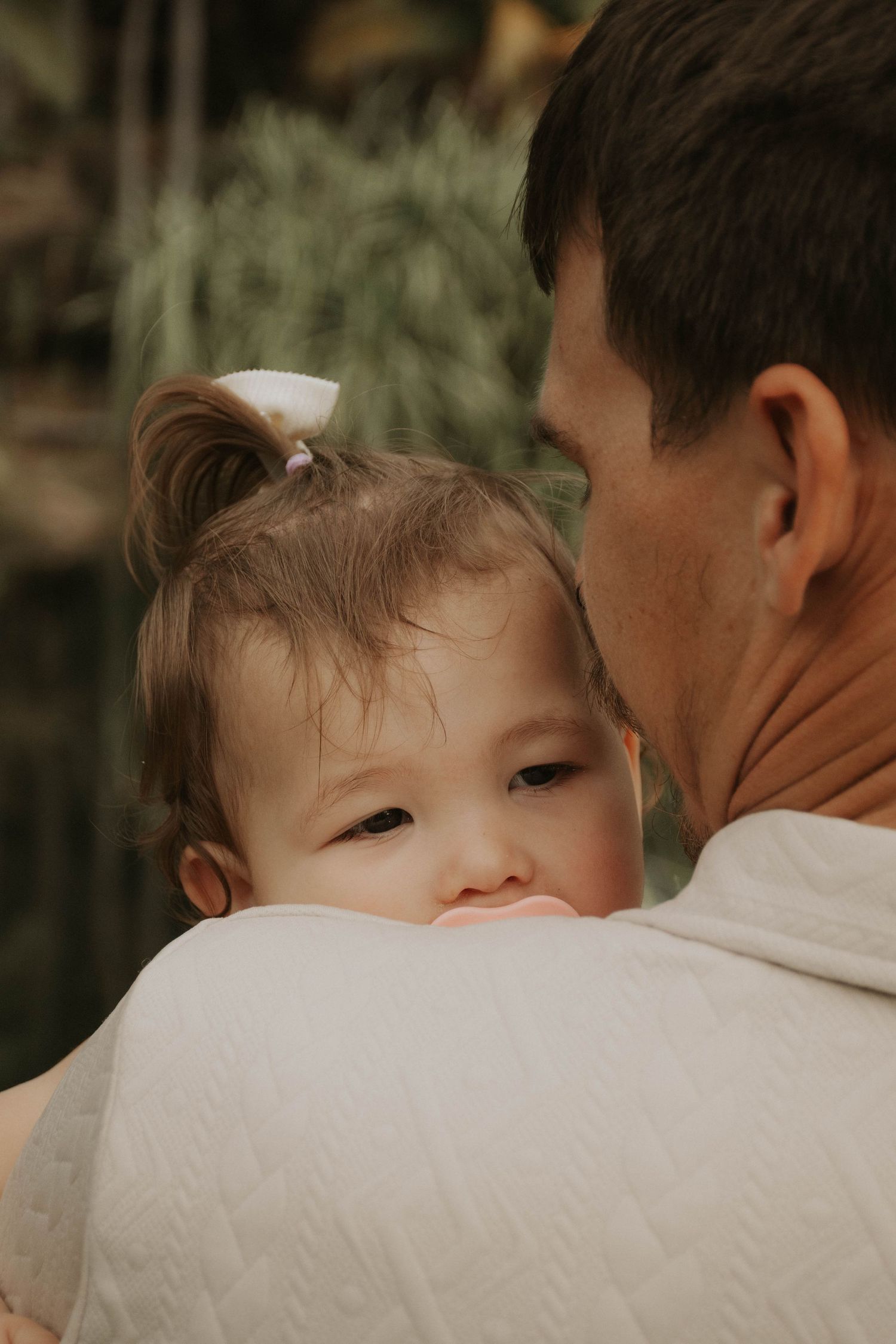 Cute baby girl with a white bow resting on fathers shoulder, gazing softly at the camera.