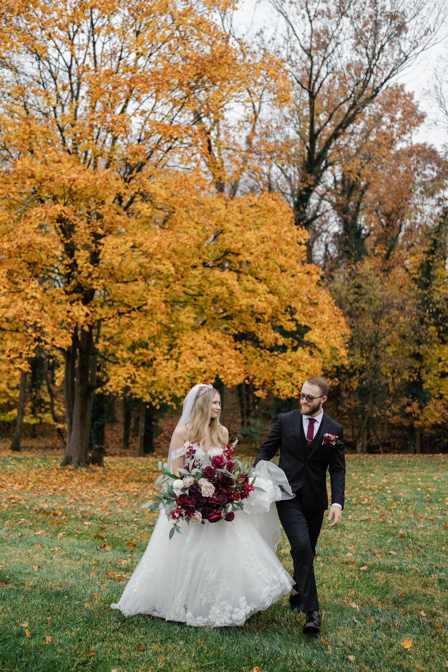 Bride and groom walking through autumn park with golden foliage, bride holding burgundy bouquet in white ball gown.